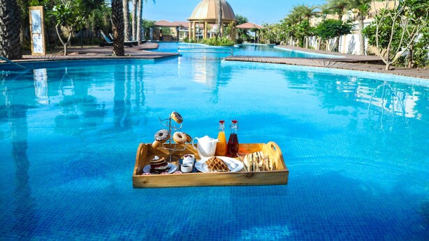Una piscina con una bandeja de madera llena de alimentos para el desayuno, como pasteles, un jarro de jugo y una botella de agua.