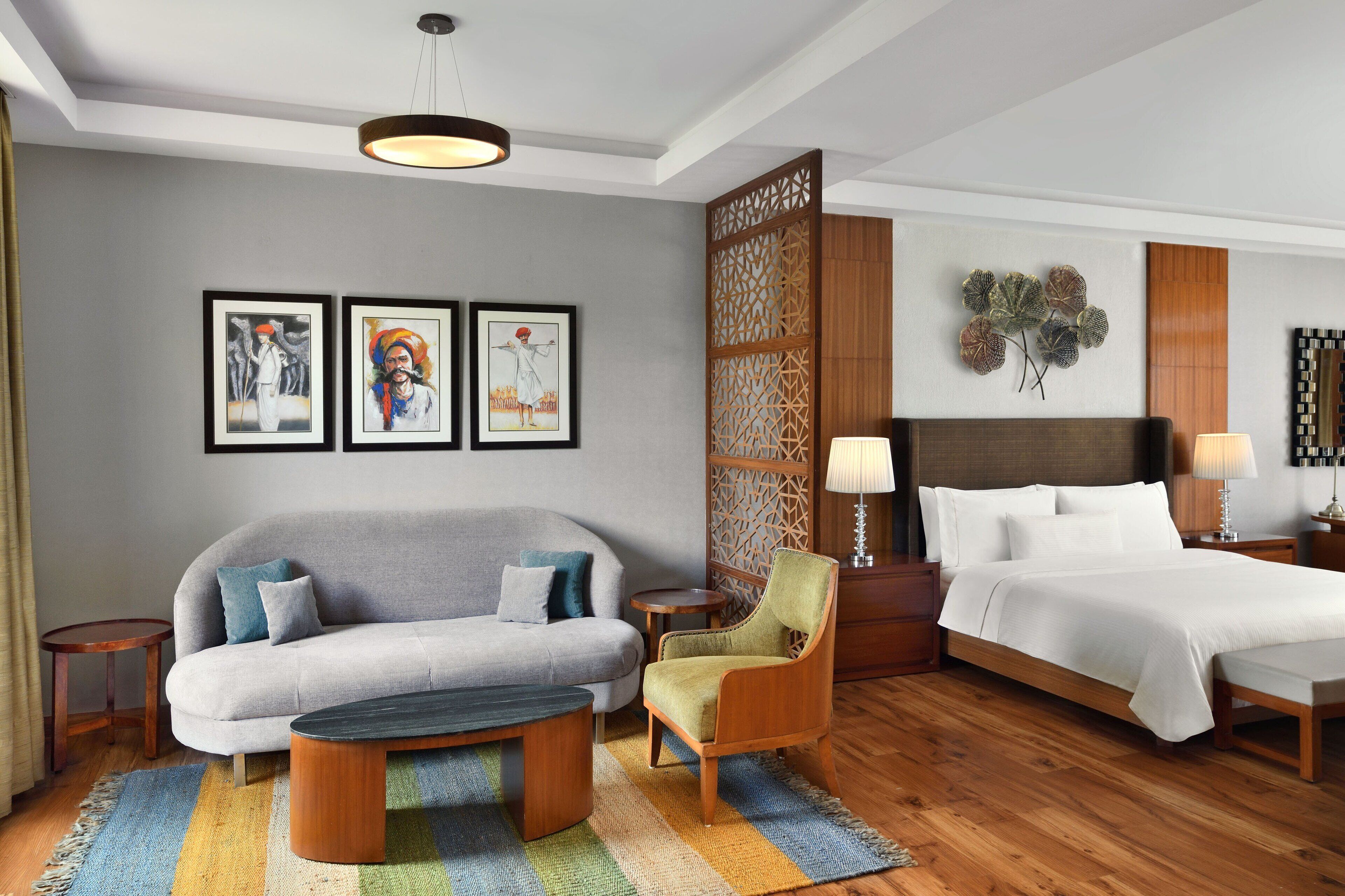 A hotel room with a sofa, armchair, and a bed. There are framed pictures on the wall, a decorative partition, and a lamp on the nightstand.