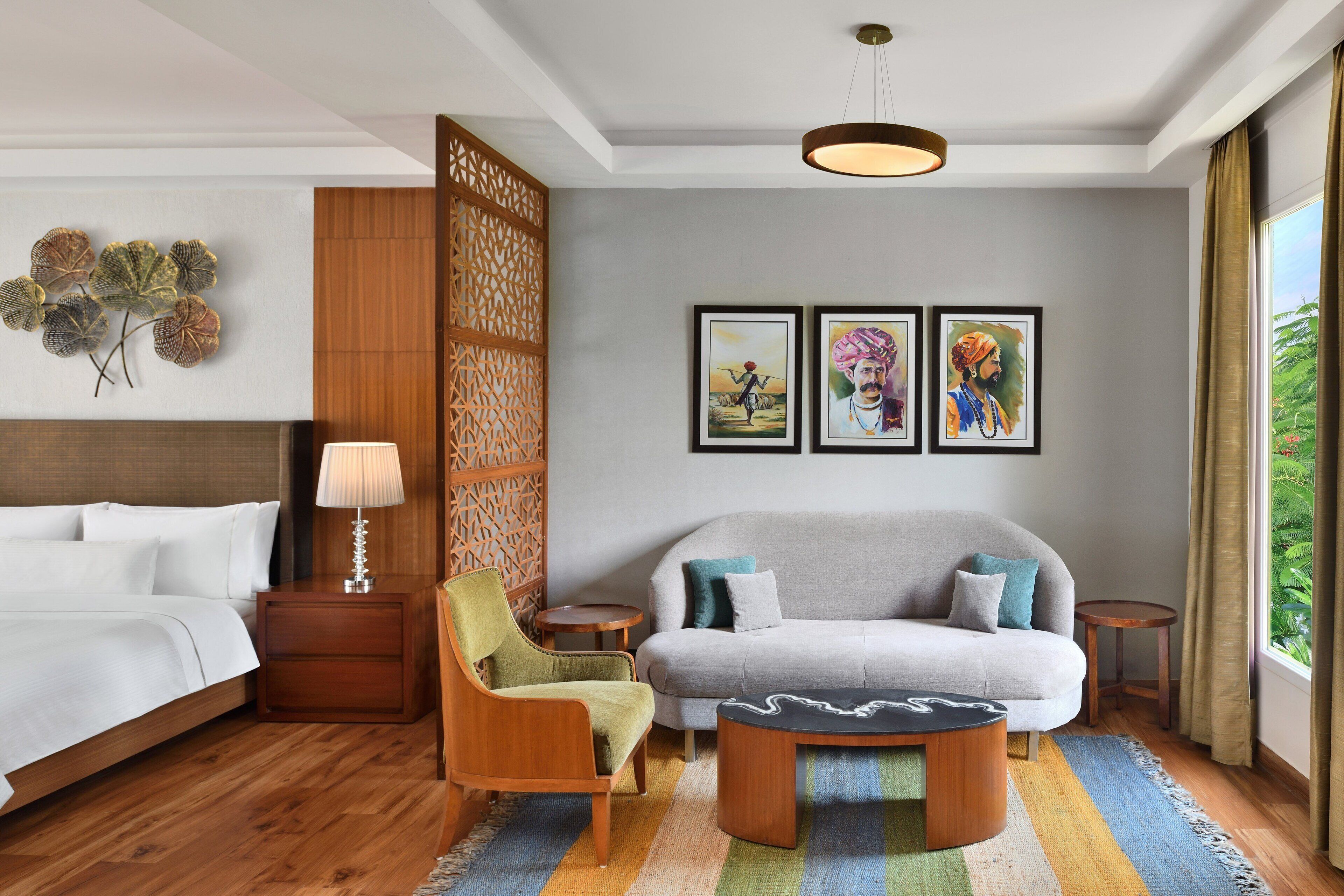 A hotel room with a bed, a sofa, a chair, a small table, a lamp, and artwork on the wall.