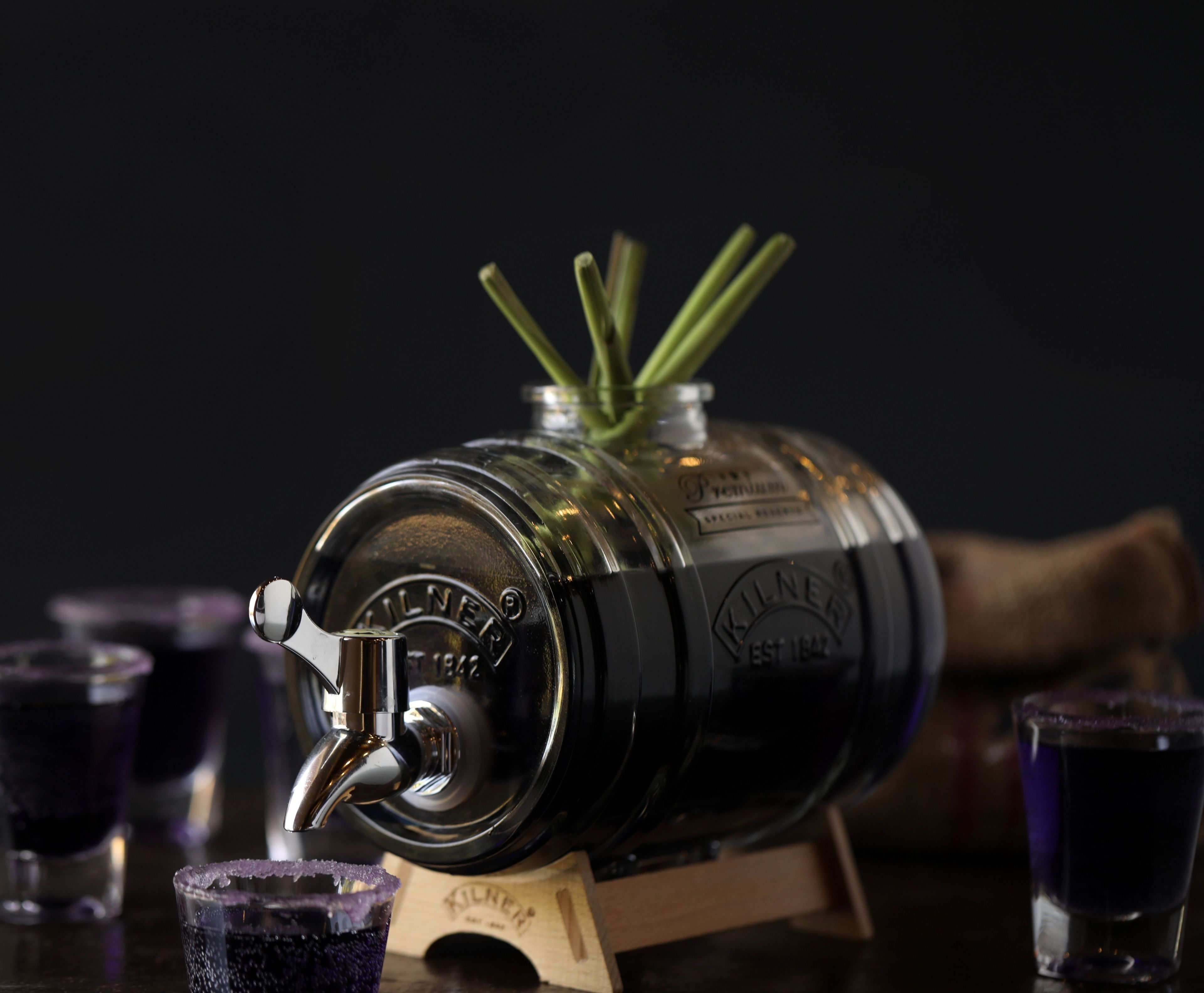 A large, black, metallic container with a tap, surrounded by glasses of dark liquid, likely a beverage.
