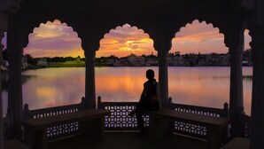 Una persona sentada en un banco bajo una estructura con arcos, contemplando un cuerpo de agua tranquilo y un atardecer.