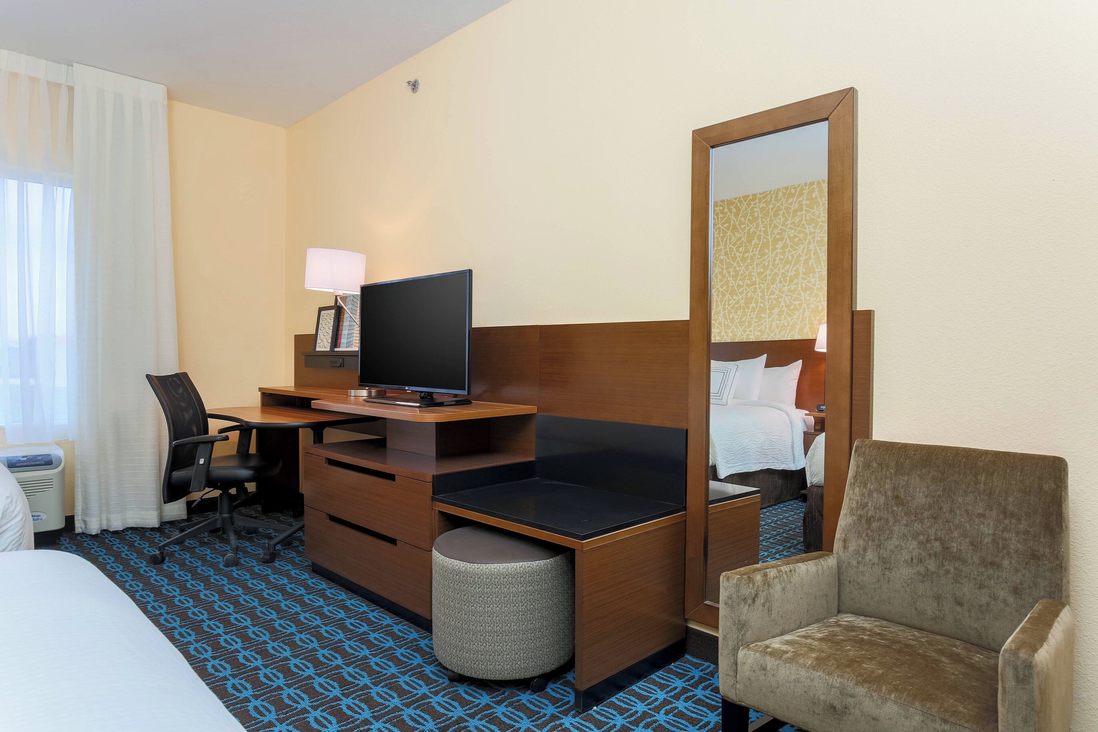 A hotel room with a bed, a desk with a computer, a chair, a small round ottoman, a mirror, and a television.