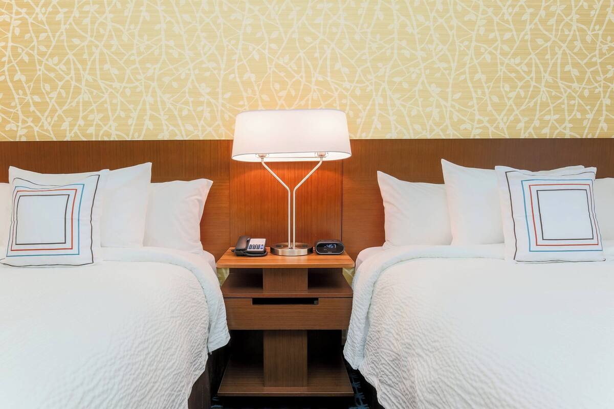 A hotel room with two beds, a nightstand with a lamp, a phone, and a clock.