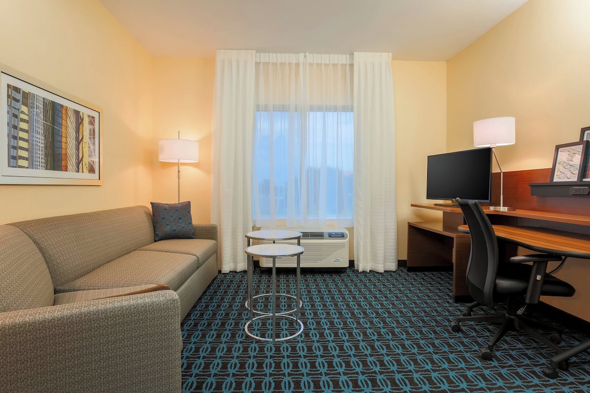 A hotel room with a sofa, a small round table, a desk with a chair, a TV, and a window with curtains.