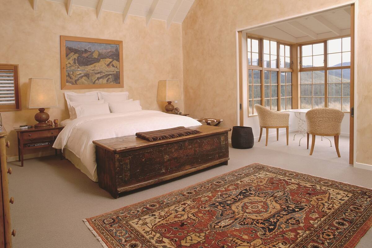 Ein Schlafzimmer mit einem großen Bett, zwei Nachttischen, einer Holzkommode, einem gemusterten Teppich, einem Korbstuhl und einem großen Fenster mit Blick.