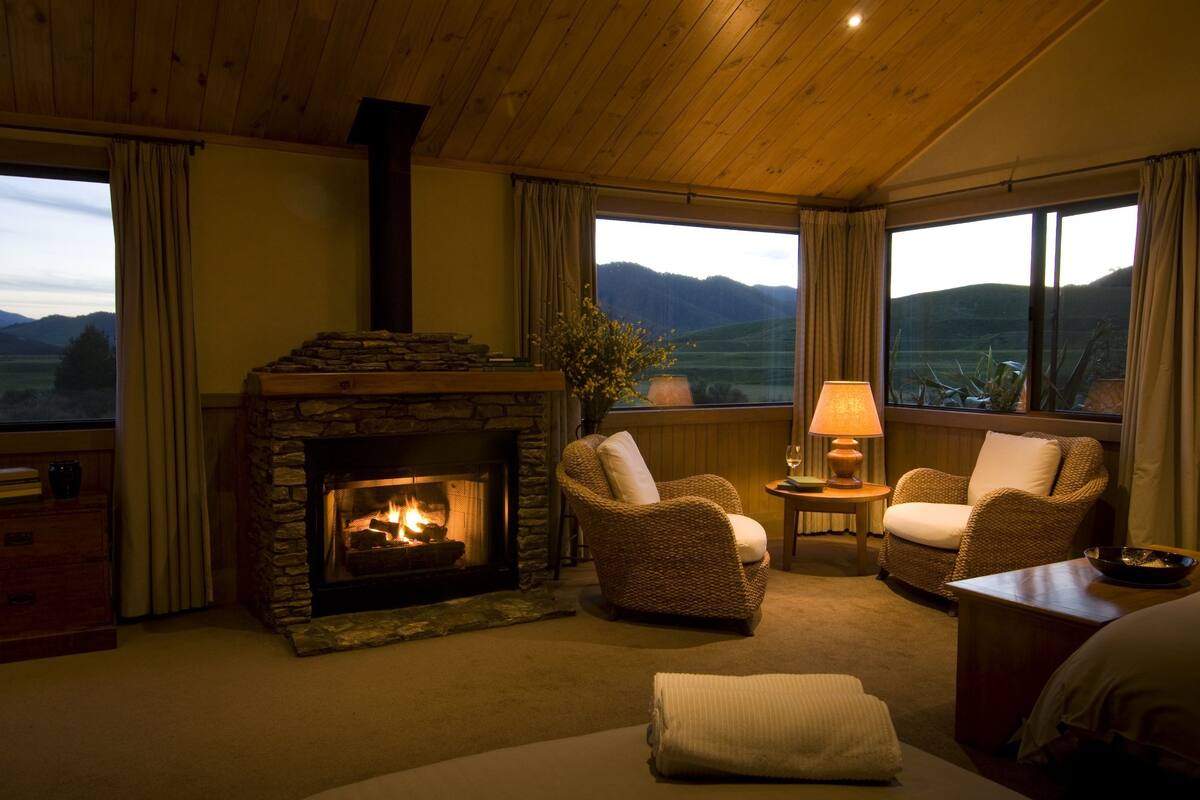 Ein gemütlicher Raum mit Kamin aus Stein, Korbsesseln, einer Lampe und großen Fenstern mit Blick auf die Berge.