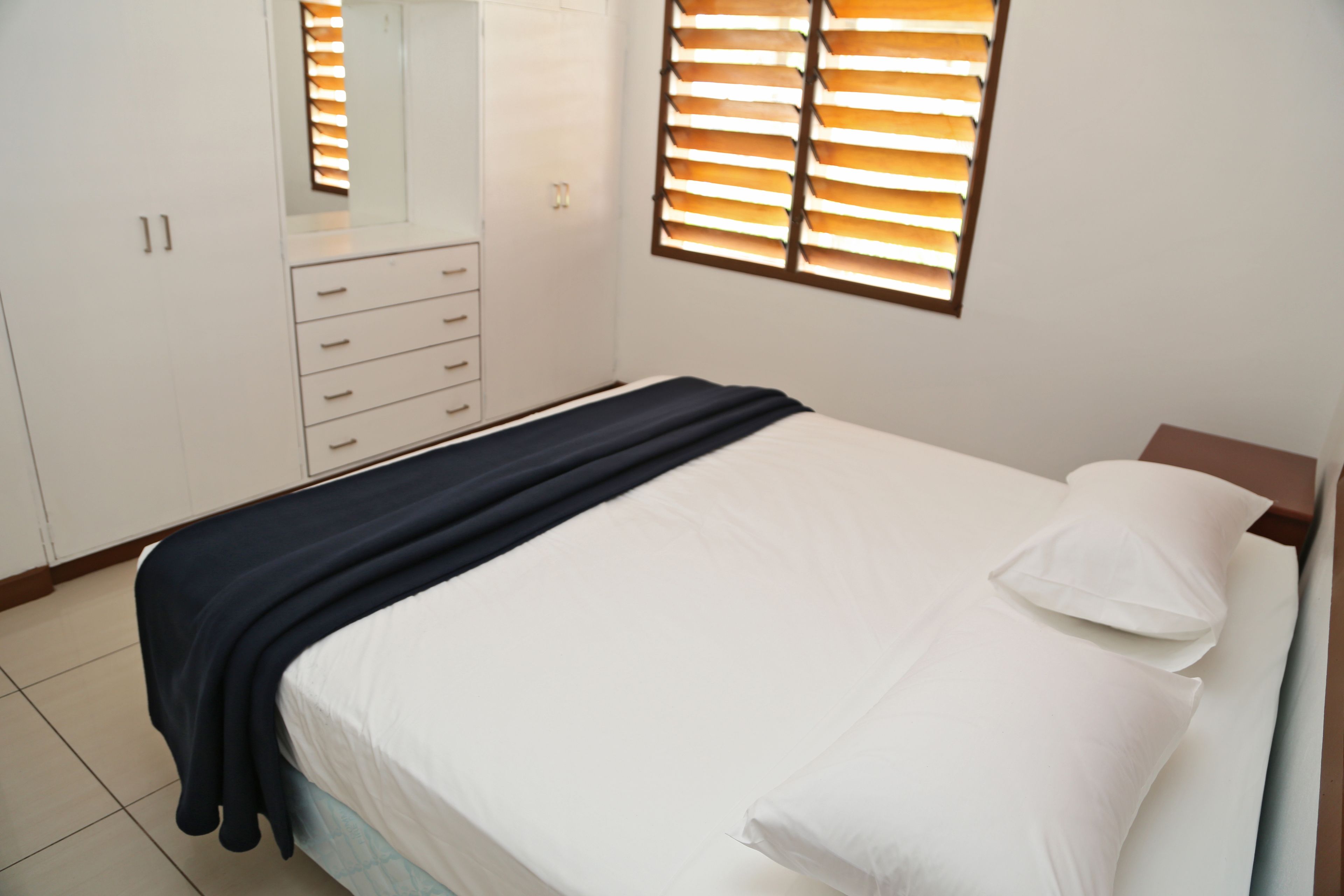 A bedroom with a bed, white bedding, a dark blue throw blanket, a wooden headboard, and a window with blinds.