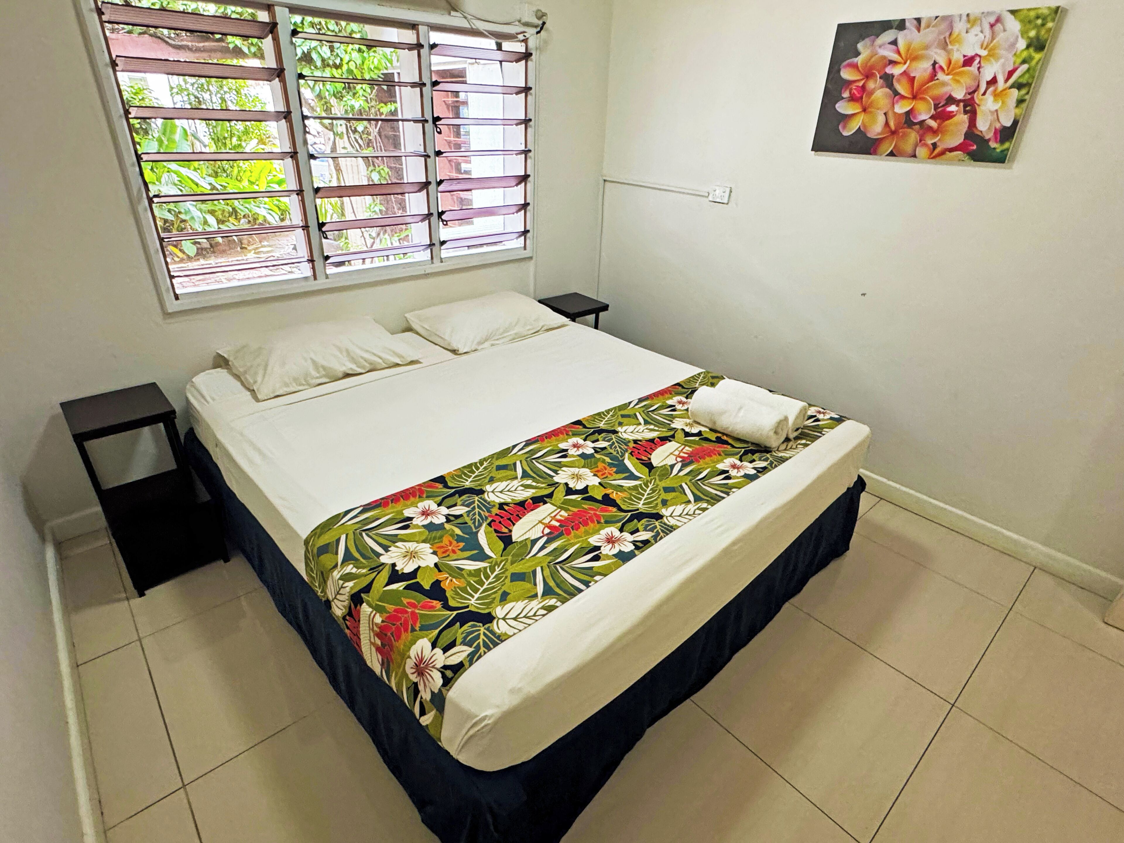A neatly made bed with a floral bedspread, a window with blinds, and a wall-mounted power outlet.