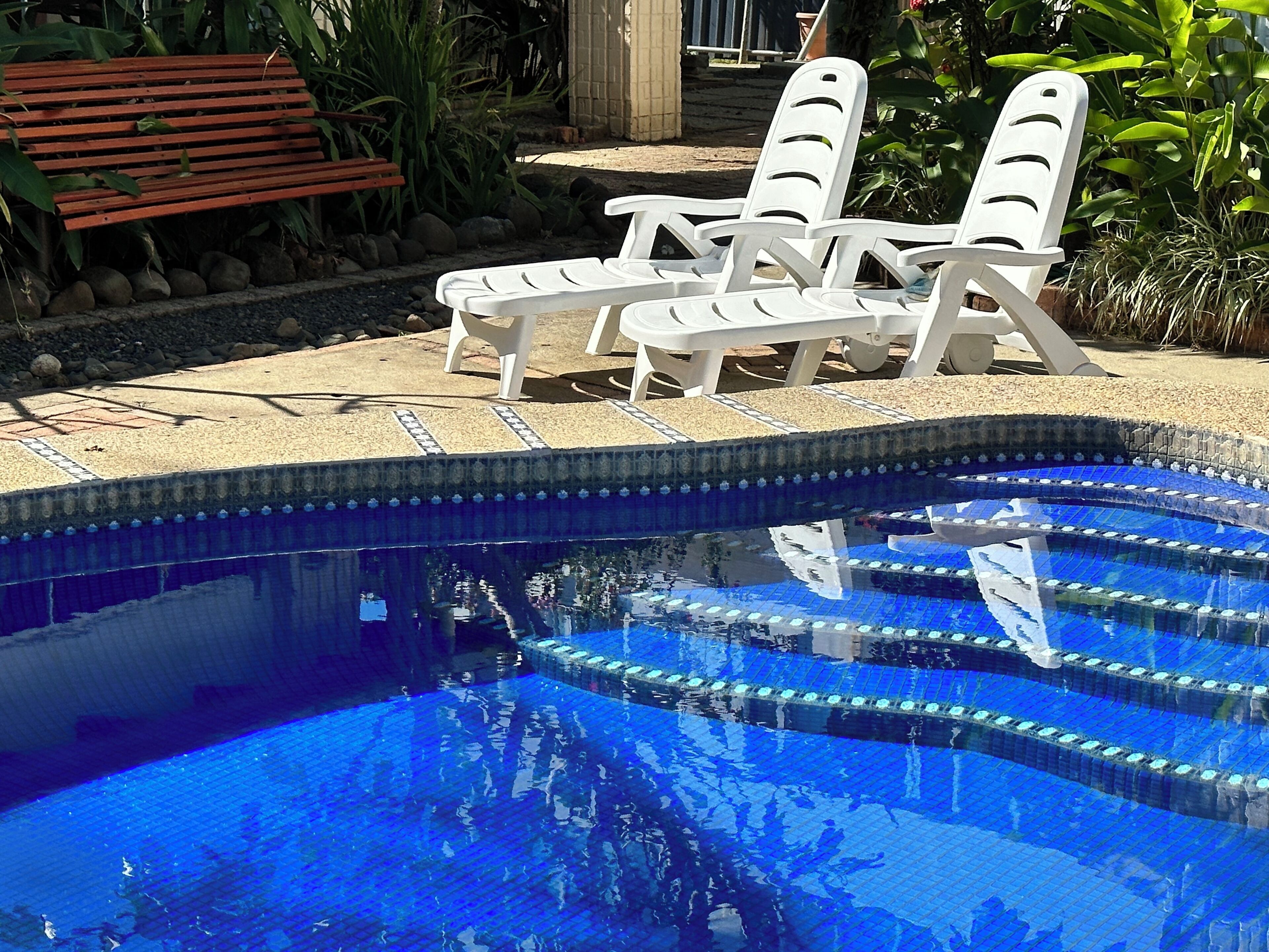 Ein Schwimmbecken mit klarem, blauem Wasser, umgeben von einer gefliesten Terrasse. Zwei weiße Liegestühle und eine Holzbank sind vorhanden.