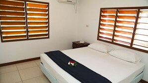 A neatly made bed with a dark blanket and a floral decoration, wooden window blinds, a small bedside table with a book, and a wall-mounted air conditioner. - Bua Bed & Breakfast (Nadi)