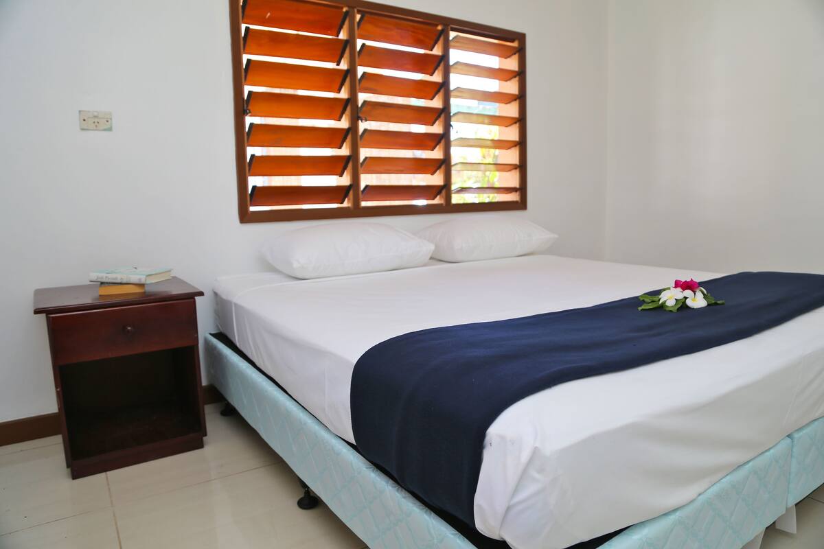 A neatly made bed with a dark blue blanket and white pillows, a wooden nightstand with a book, and a window with wooden blinds.