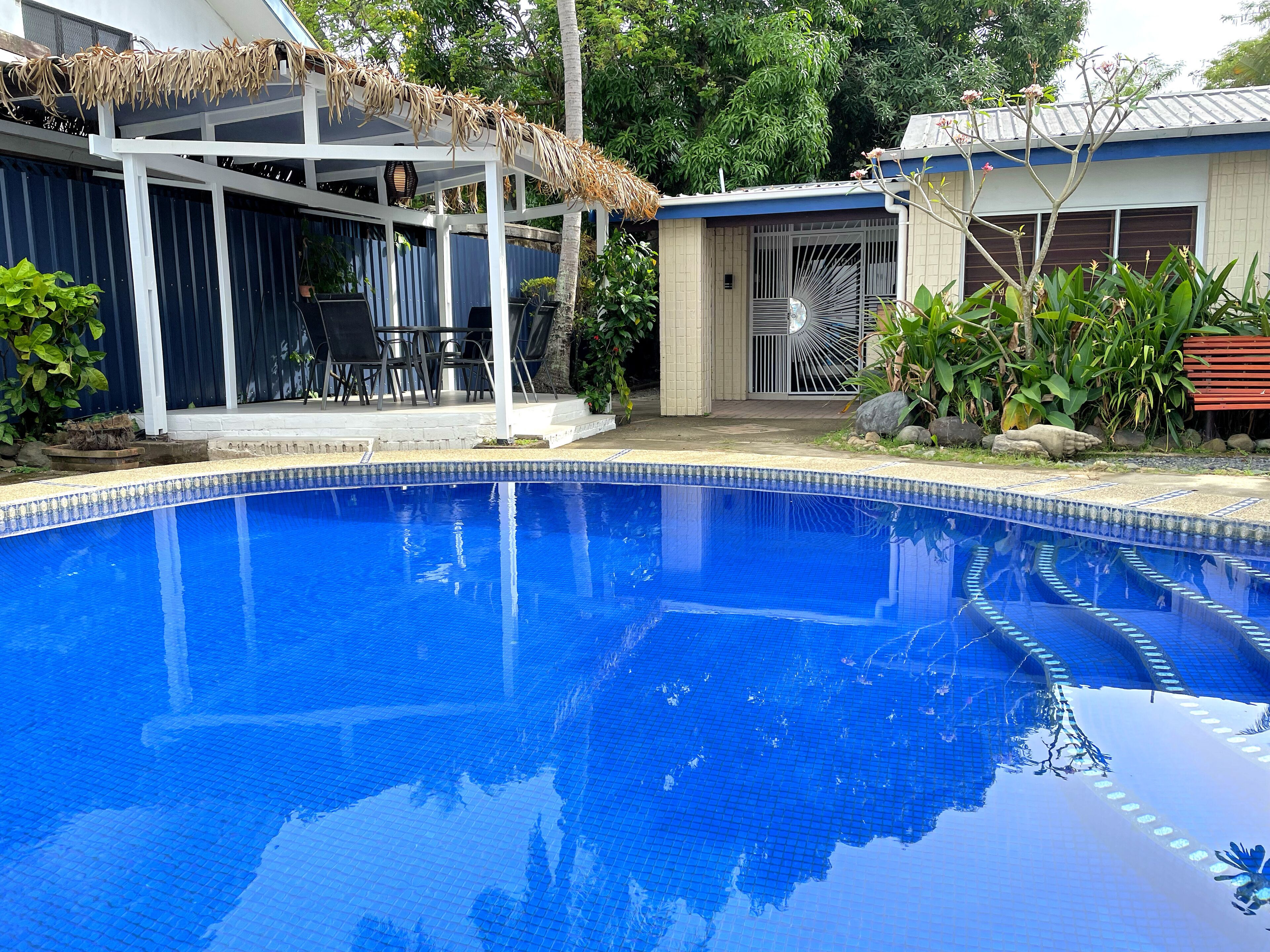 A swimming pool with a tiled edge, surrounded by a covered patio area with chairs and a table.