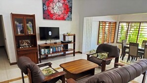 A living room with a sofa, armchairs, a coffee table, a TV, a bookshelf, and a window with a view of greenery. - Bua Bed & Breakfast (Nadi)