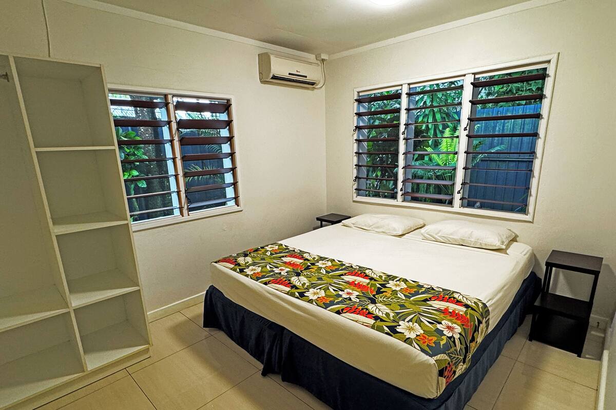 A bedroom with a bed, two windows with shutters, a wardrobe, and an air conditioning unit.