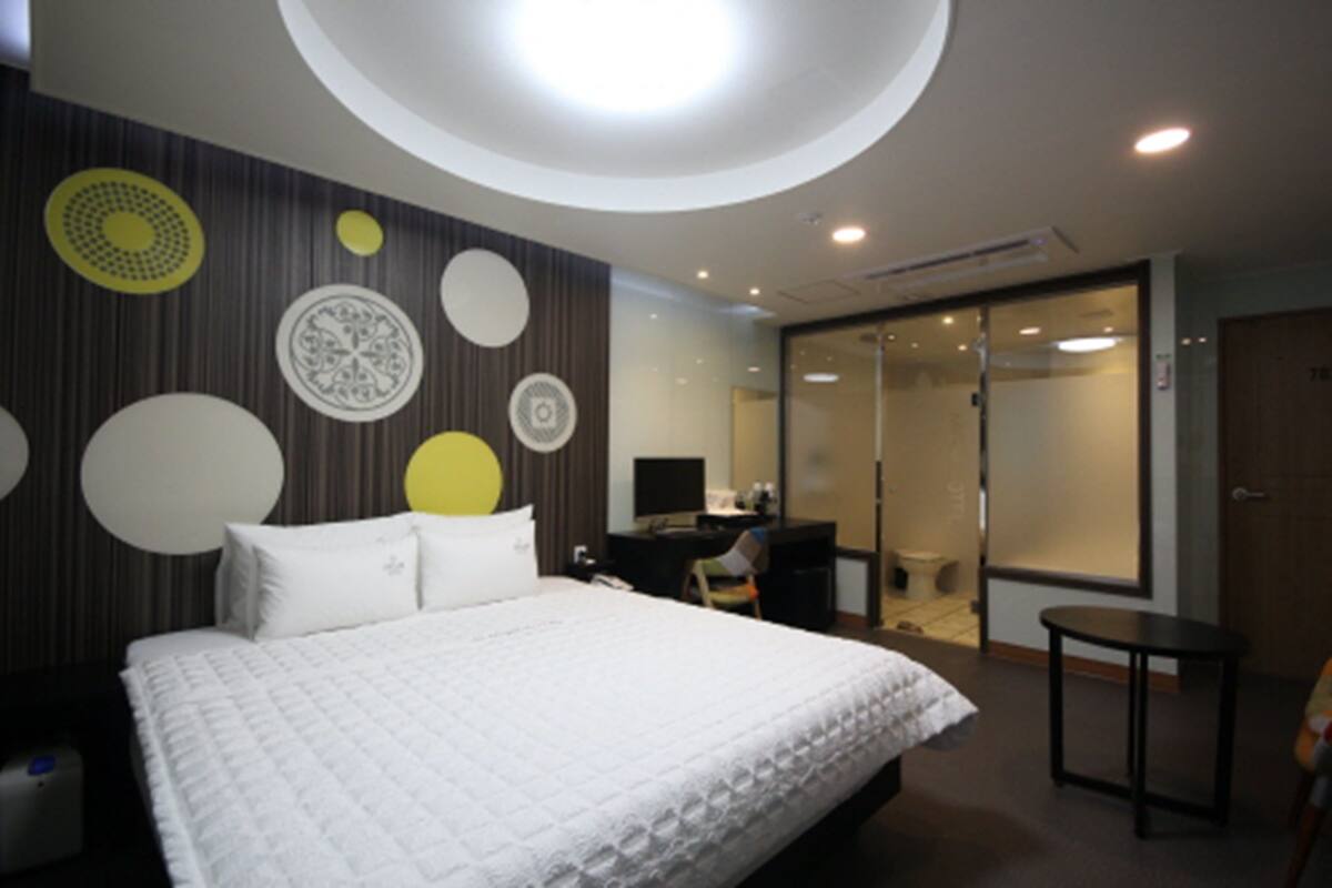 A modern hotel room with a large bed, a desk, and a small table. There is a sliding door leading to a bathroom.