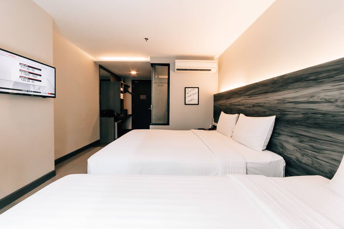 A hotel room with two beds, a flat-screen TV, an air conditioning unit, and a framed quote on the wall.