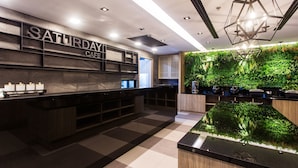 A modern cafe interior with a checkered floor, a wooden reception desk, a wall-mounted plant display, and a geometric ceiling light fixture. - Spenza Hotel (Bangkok)
