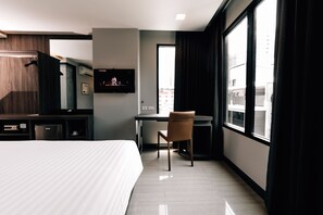 Habitación de hotel moderna con cama, escritorio, silla, televisor y ventanas amplias con vista a la ciudad.