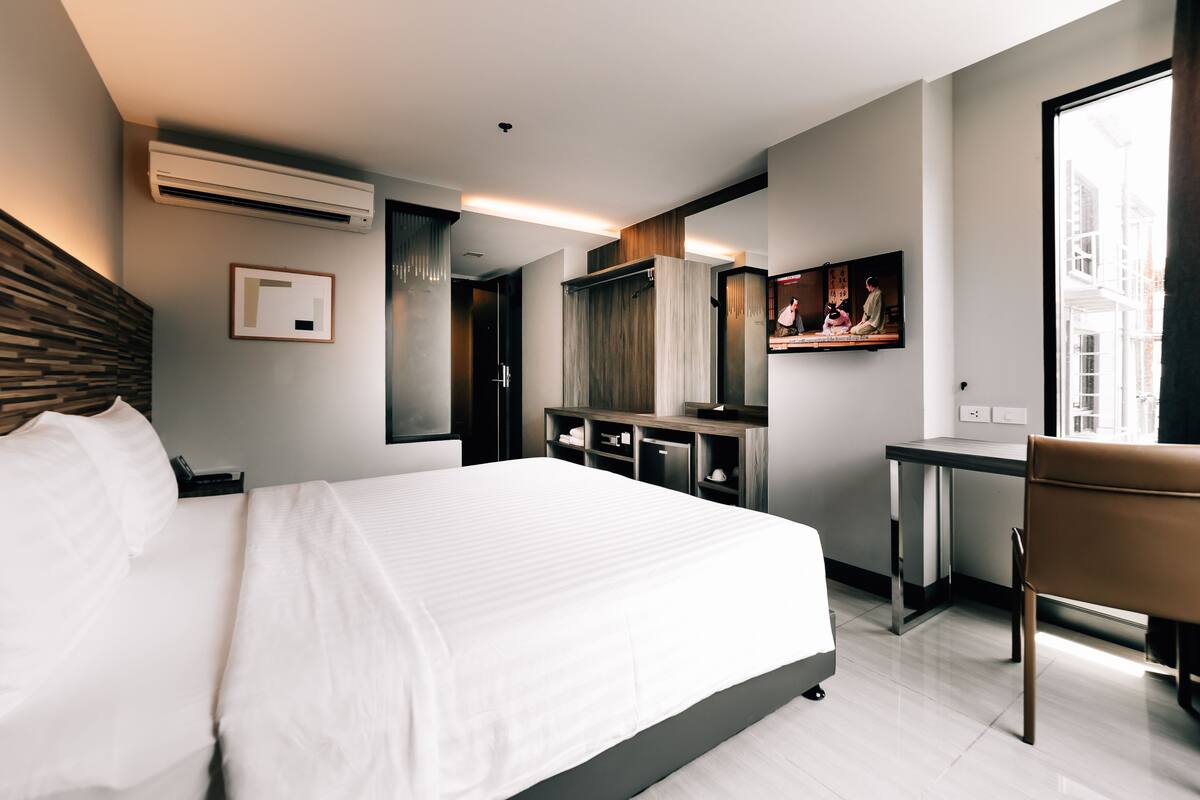 A modern hotel room with a large bed, a desk, and a chair. There is a mirror, a TV, and an air conditioning unit on the wall.