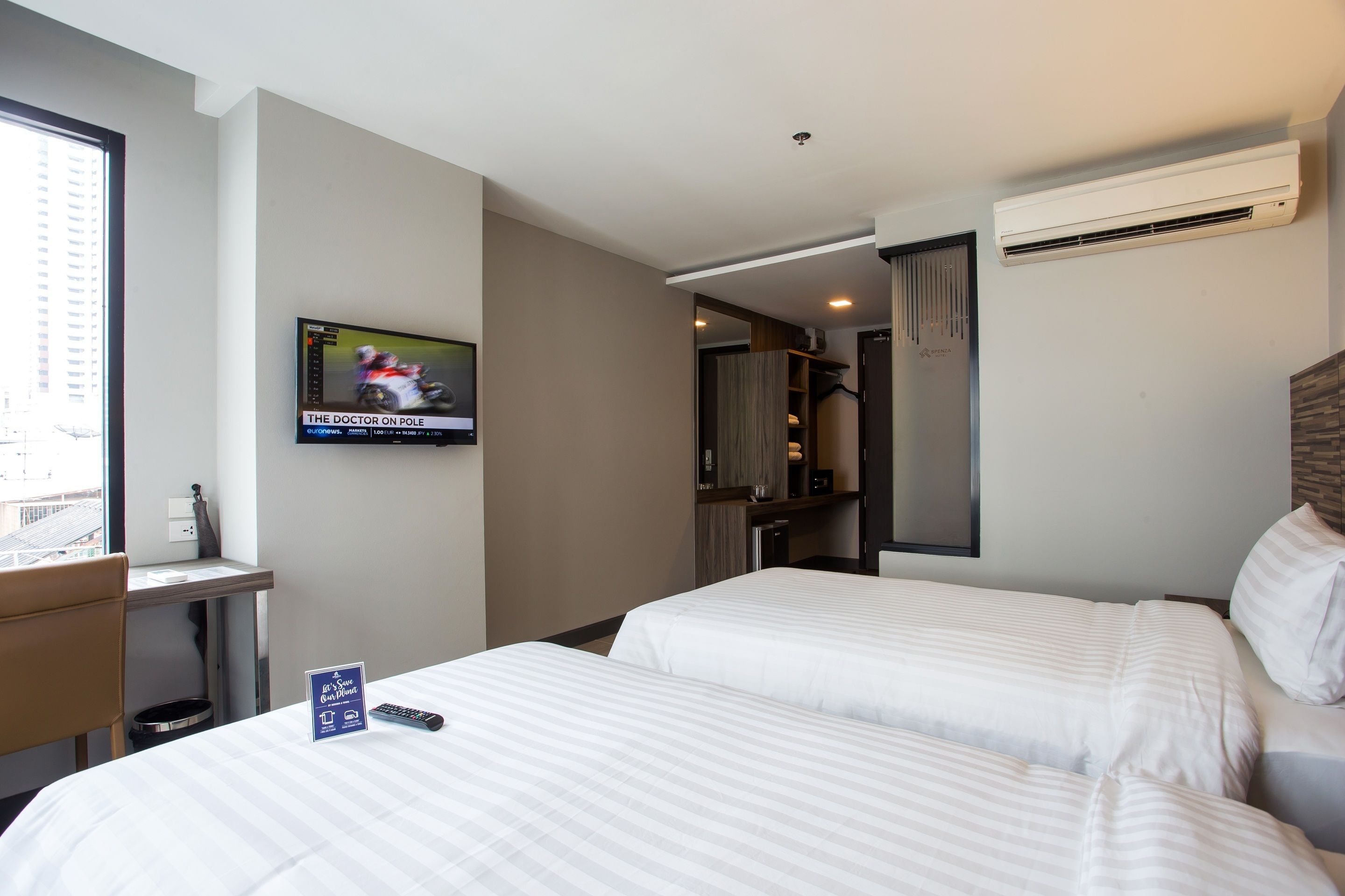 A hotel room with two beds, a desk, a TV displaying a racing game, and an air conditioning unit.