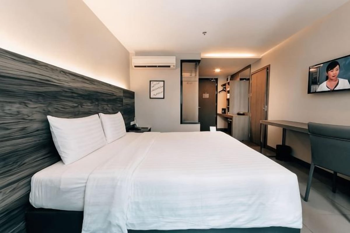 A hotel room with a large bed, a desk, a chair, a TV, and a wardrobe.
