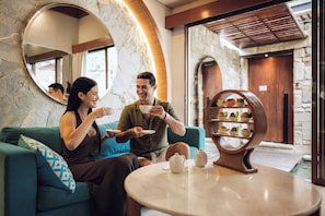 A man and a woman sitting on a teal sofa, holding plates and cups, with a coffee table in front of them.