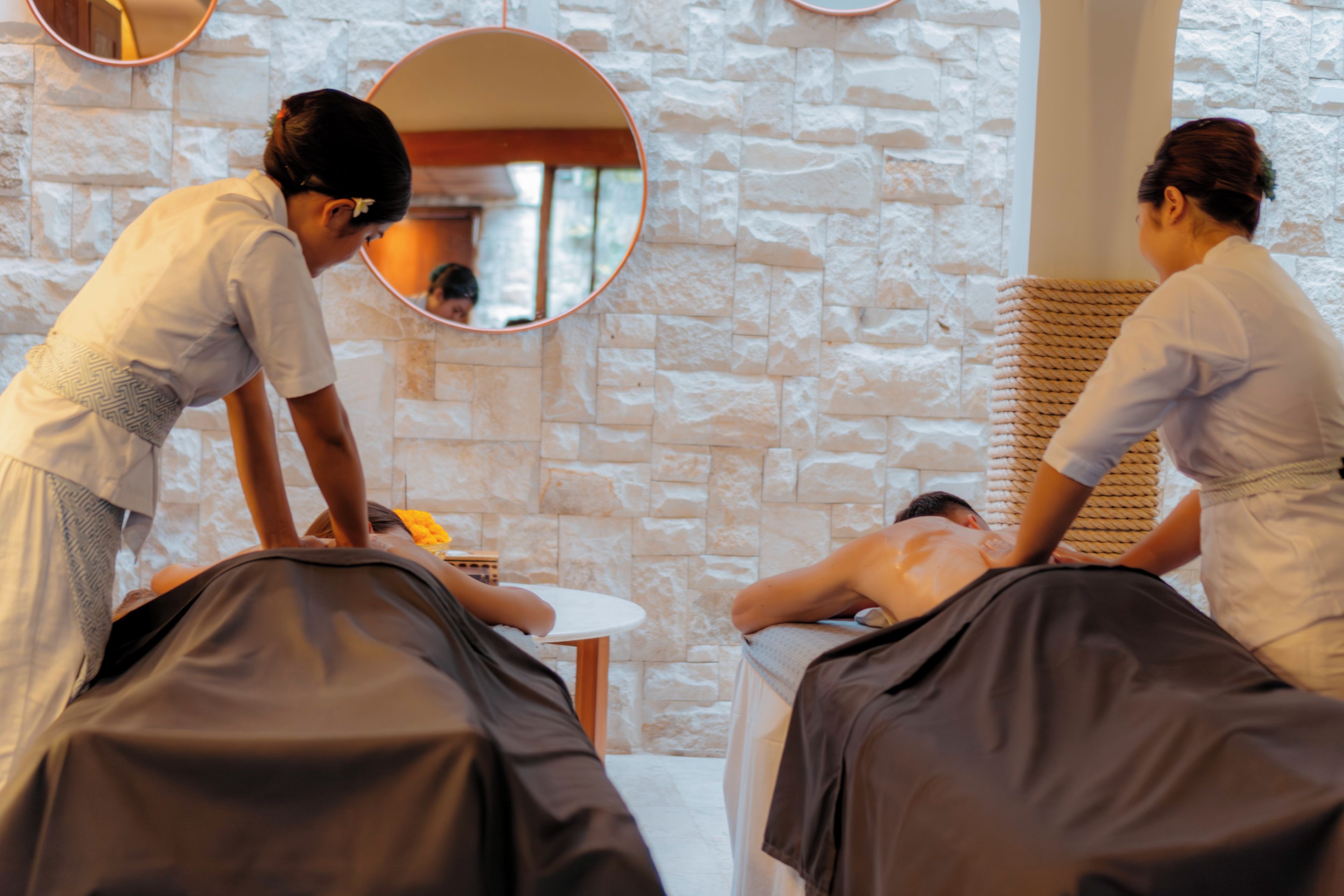 A massage therapy room with two massage tables, a stone wall, and a mirror.