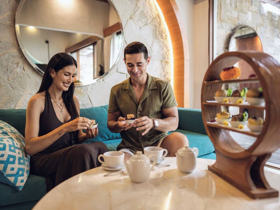 A couple sitting on a sofa with a table in front of them, a mirror on the wall, and a dessert display.