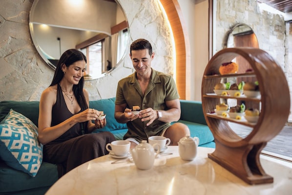 A couple sitting on a sofa with a table in front of them, a mirror on the wall, and a dessert display.