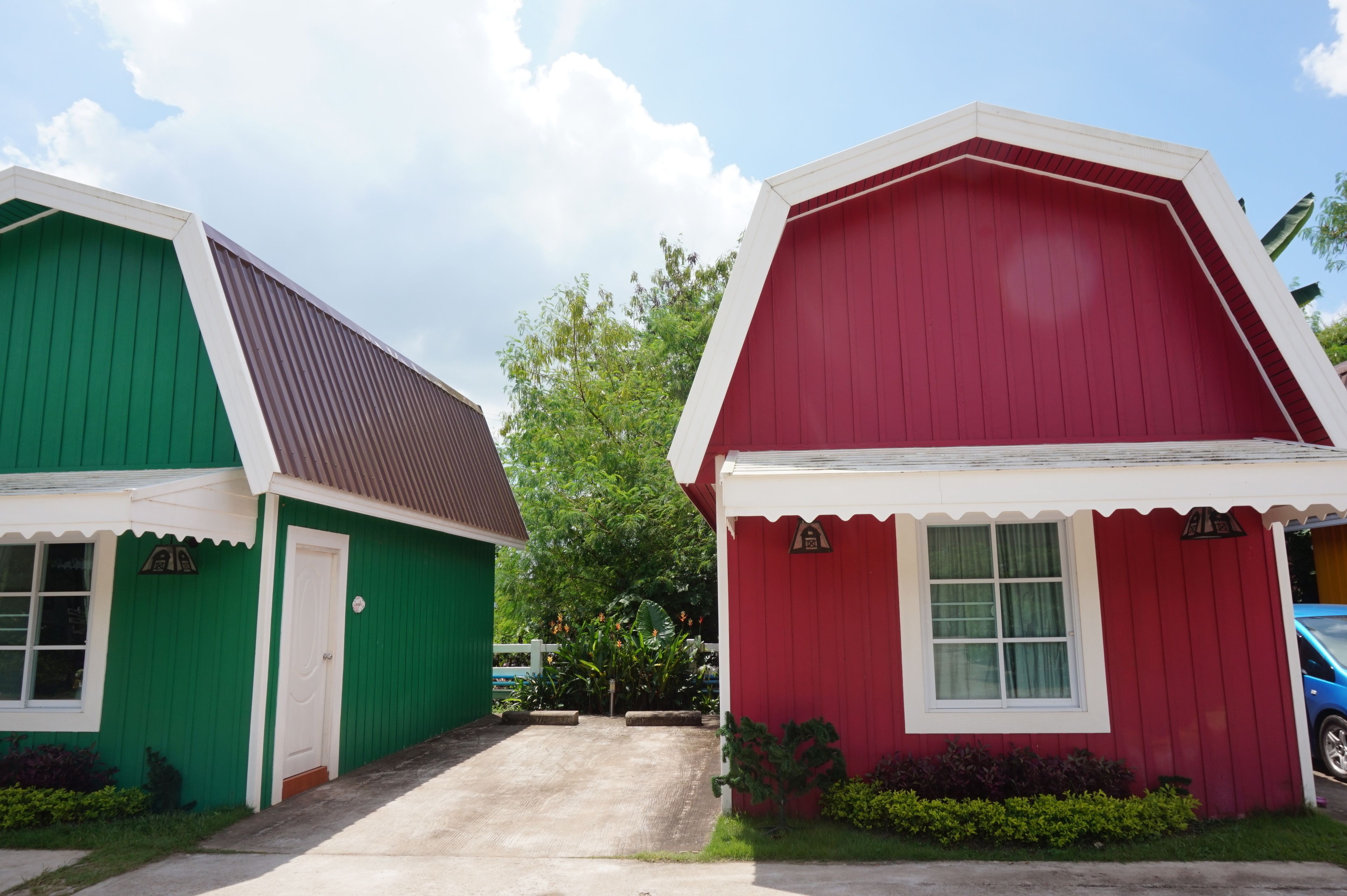 Deux petites maisons à un seul étage, aux façades vertes et rouges, avec une allée pavée et une clôture blanche en arrière-plan.