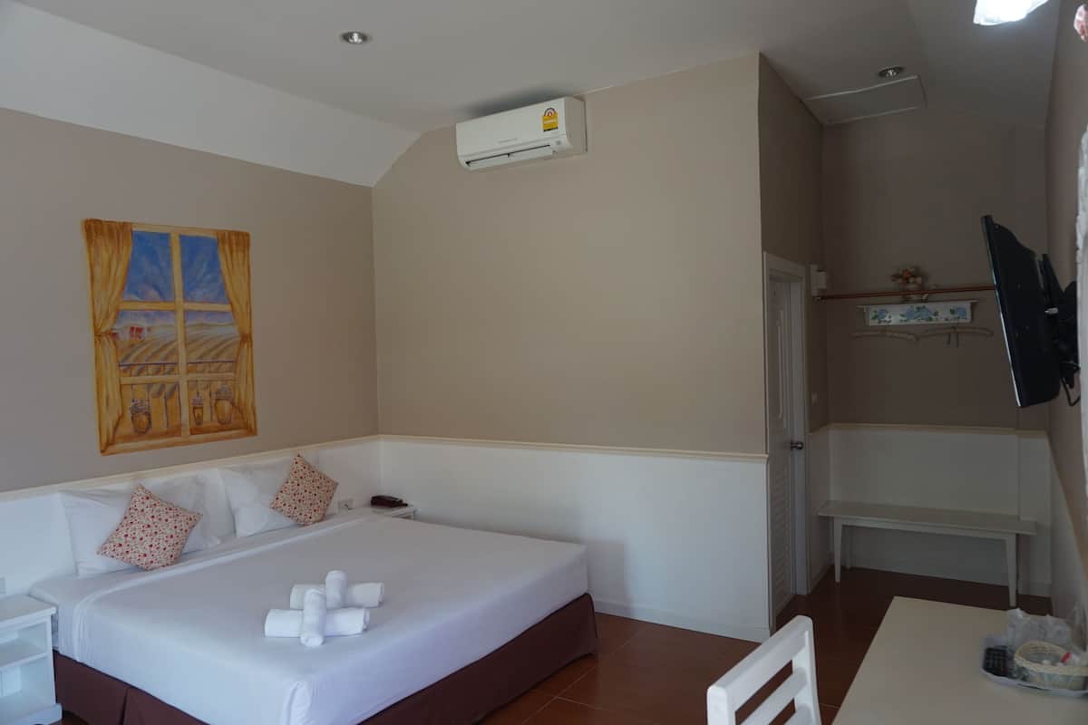 A hotel room with a bed, a small bedside table, a wall-mounted air conditioner, a painting, and a bench in the corner.