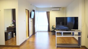 A modern living room with a flat-screen TV on a white entertainment unit, wooden flooring, a large mirror, and a sliding glass door leading to a balcony. - Suksomboon Hotel (Surat Thani)