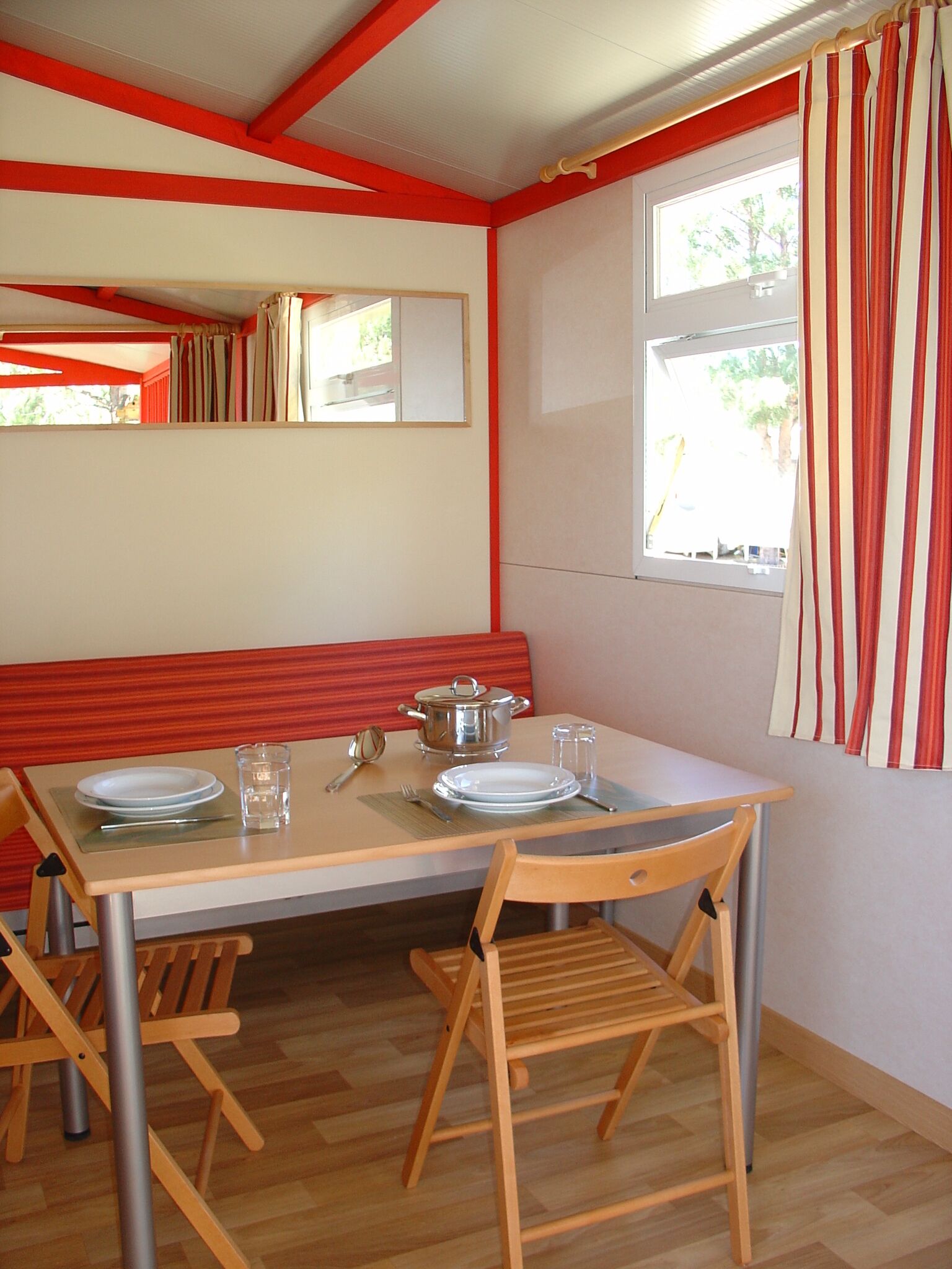 A dining area with a table set for two, wooden chairs, a bench, and a window with curtains.