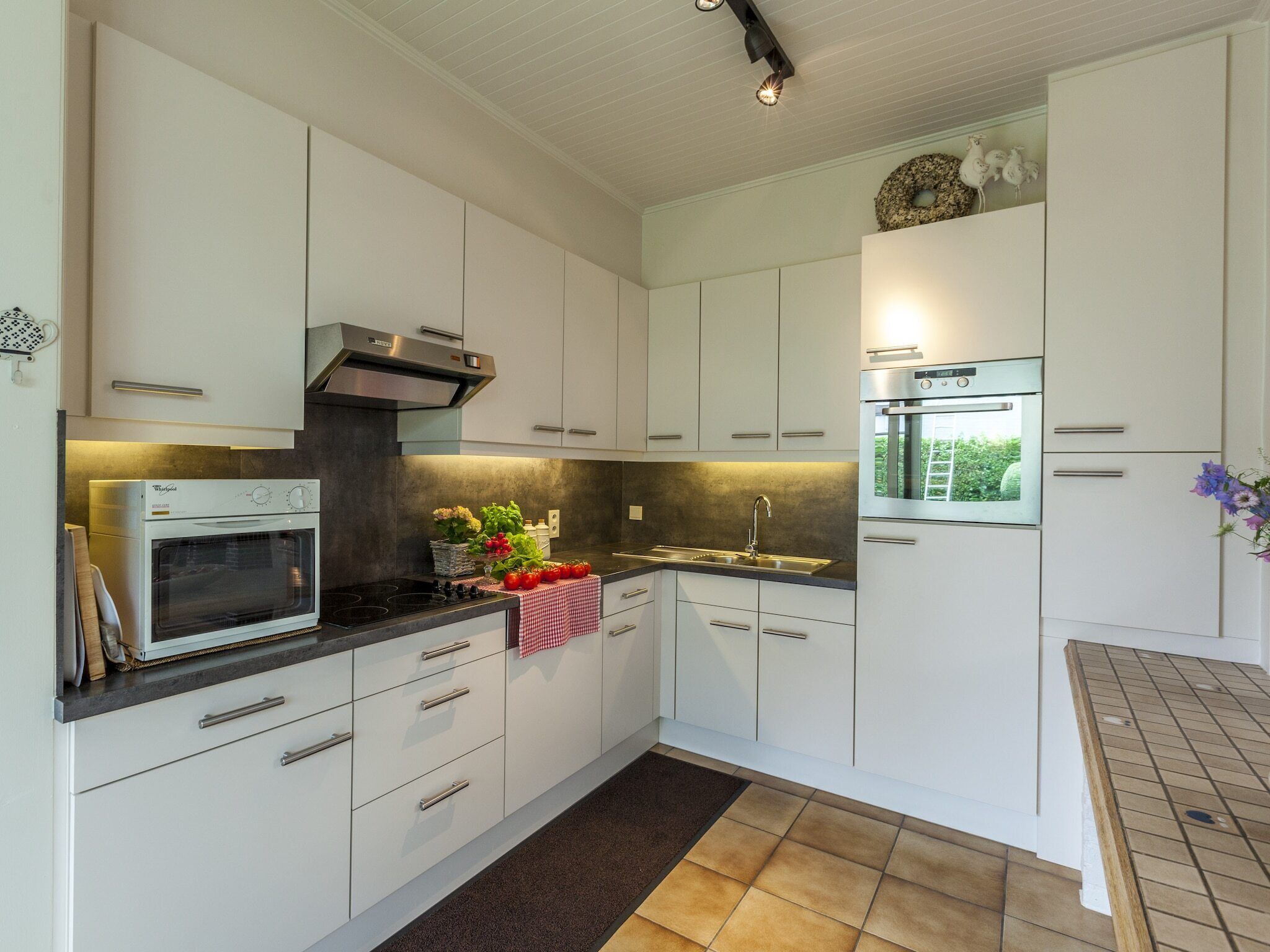 A modern kitchen with white cabinets, a built-in oven, a stainless steel range hood, and a large window.