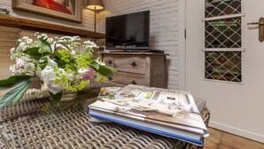 Una habitación con una mesa de mimbre, una revista, un florero con flores, un televisor sobre un mueble y una puerta con un ventanal.