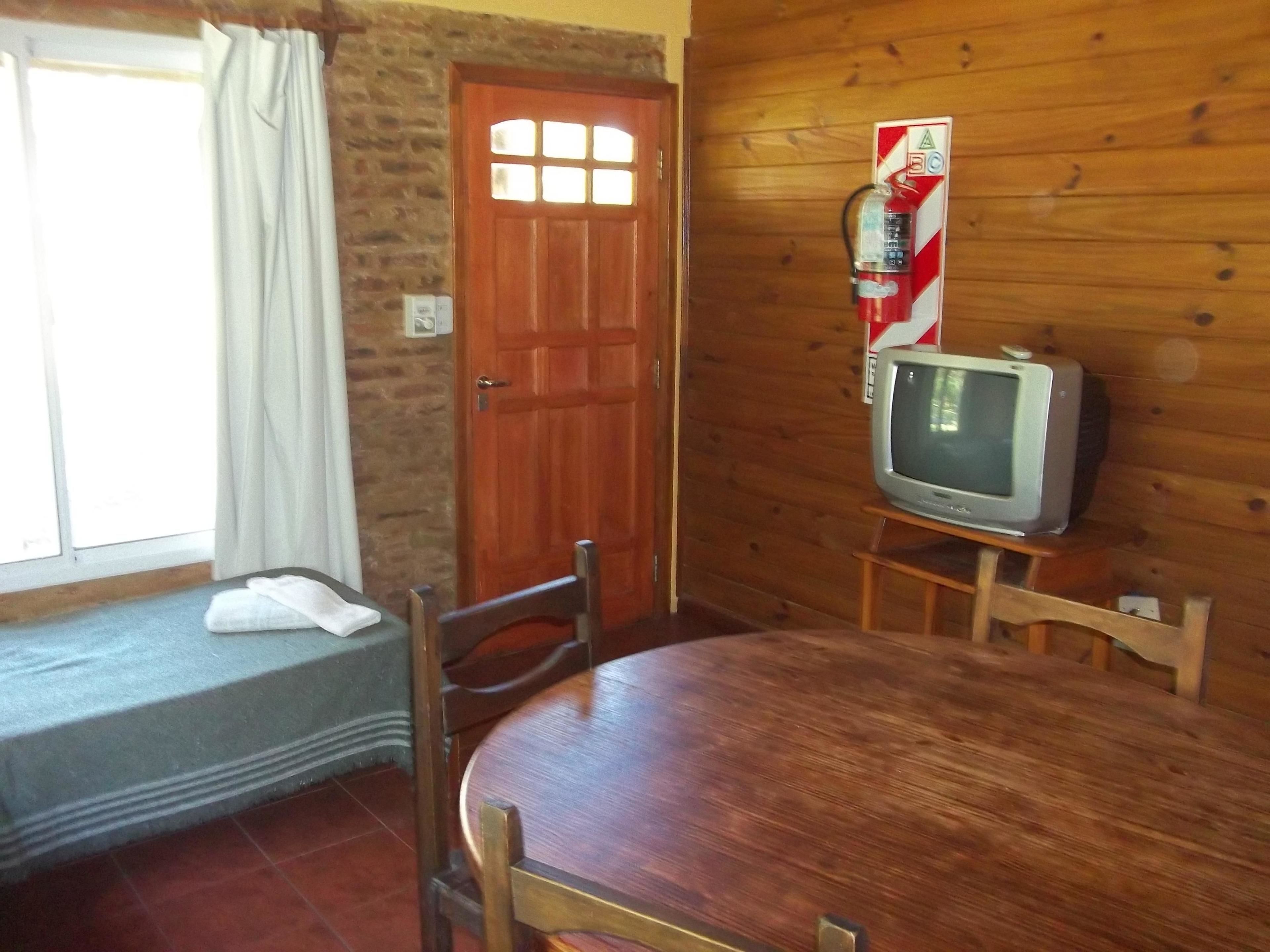 A wooden-paneled room with a round dining table, two chairs, a television on a stand, a fire extinguisher, and a window with curtains.