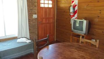 A wooden-paneled room with a round dining table, two chairs, a television on a stand, a fire extinguisher, and a window with curtains.
