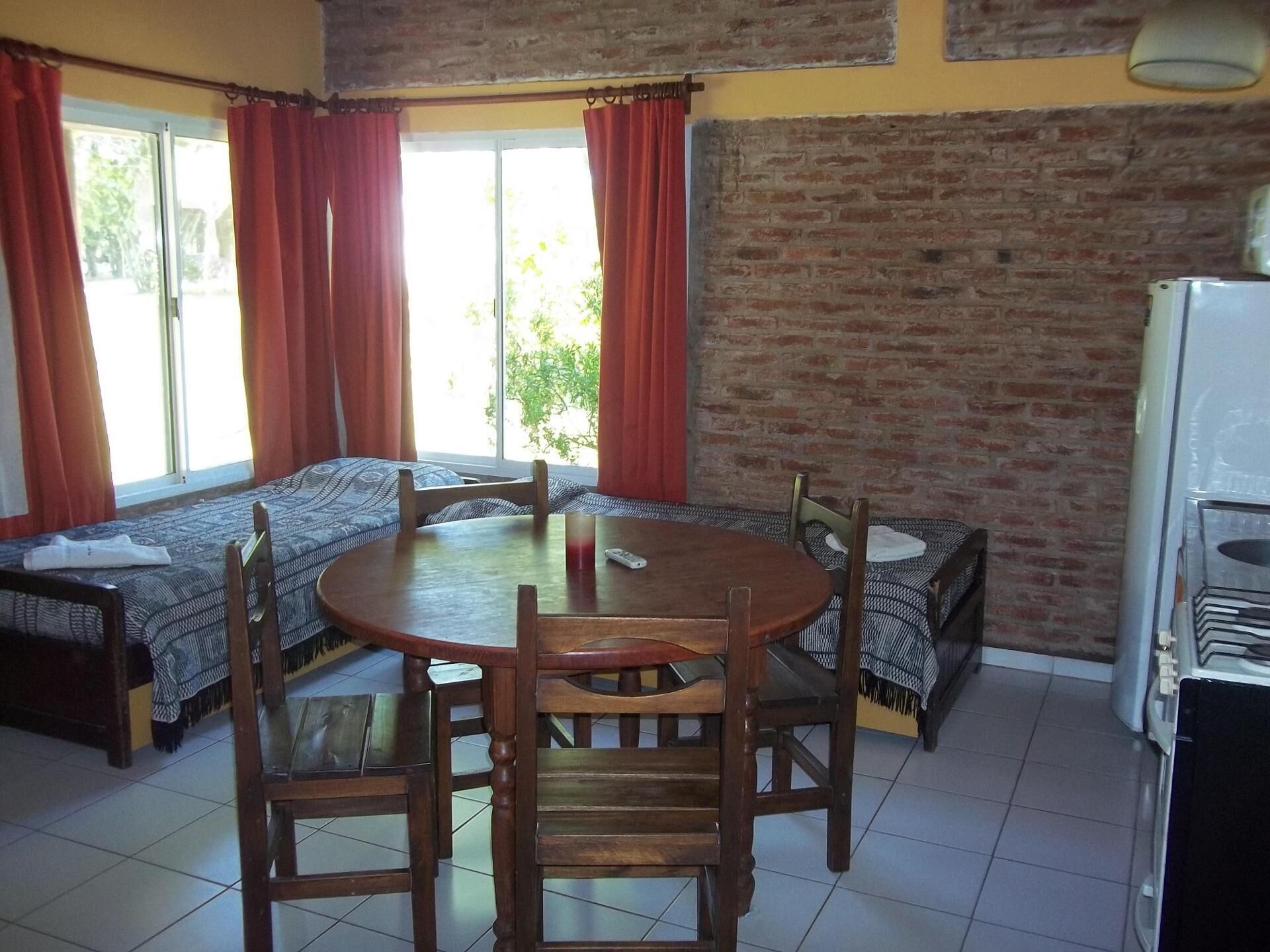 A compact kitchen with a round wooden table, four chairs, a refrigerator, and a brick wall.
