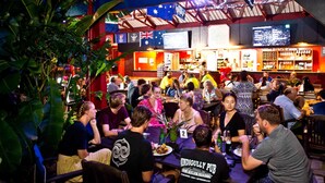 Un bar bondé, avec des tables et des chaises, un comptoir où est affiché le menu, et des drapeaux australiens.