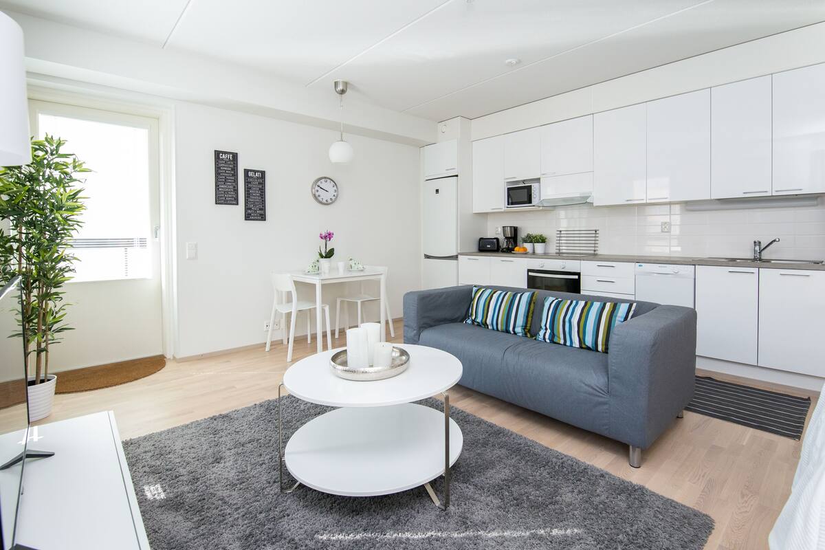 A modern apartment with a grey sofa, white dining table, and a kitchen area.