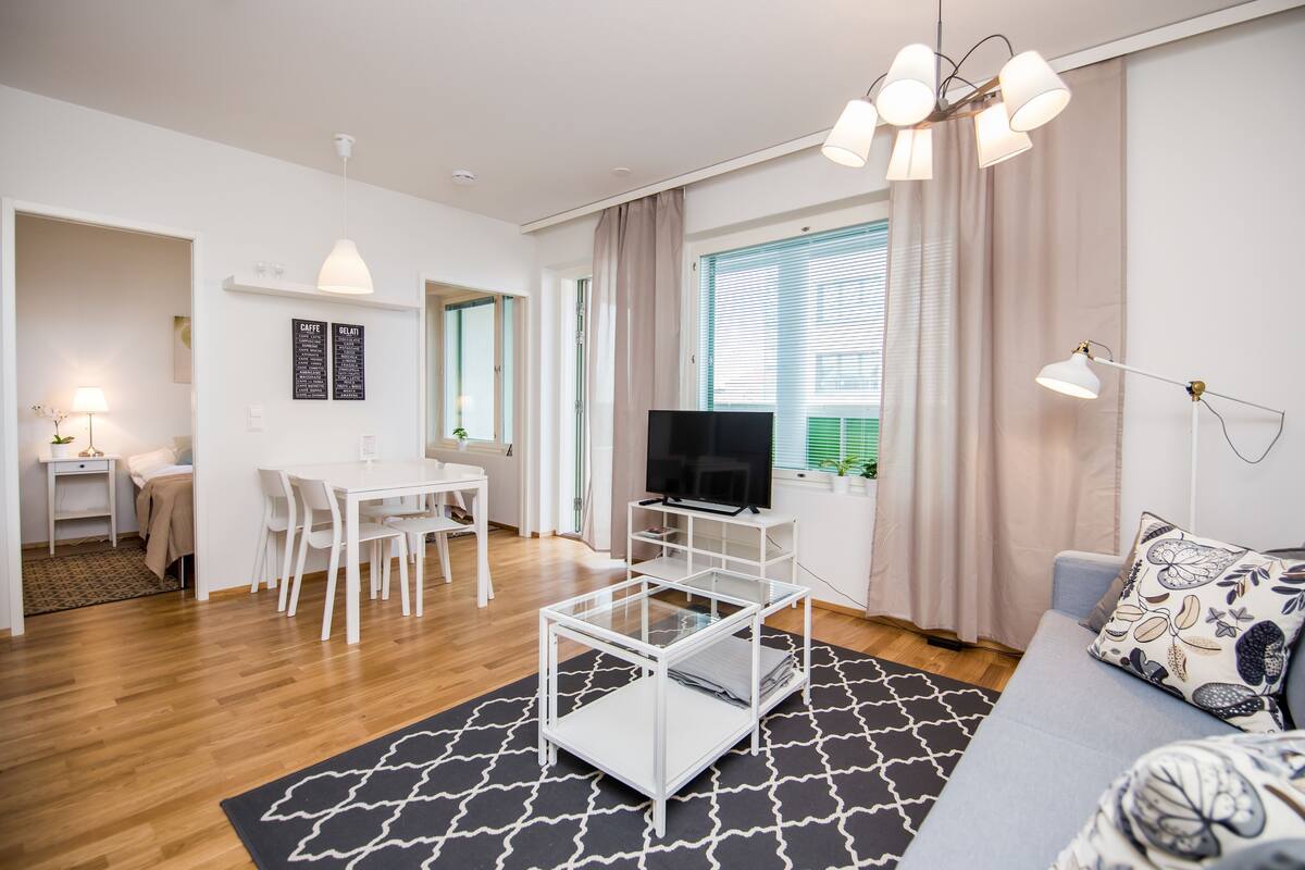 A modern living room with a glass coffee table, a flat-screen TV, a sofa with patterned cushions, and a dining area with a white table and chairs.