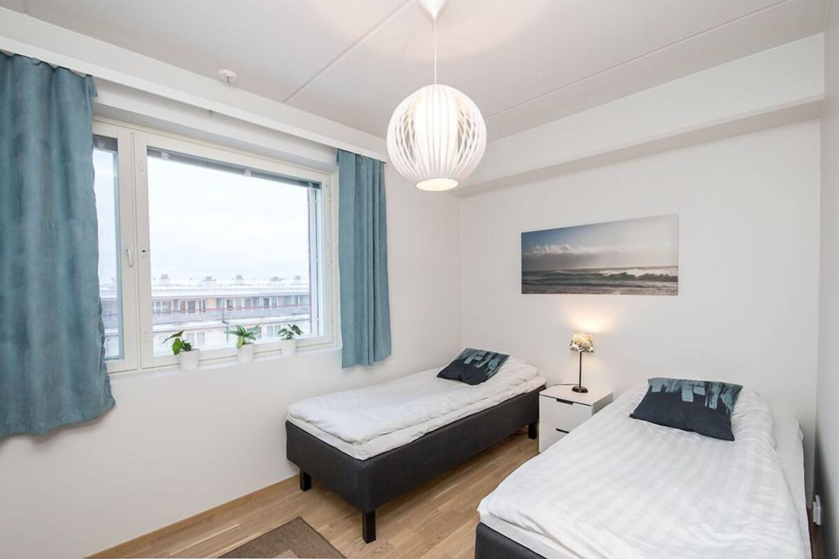 A bedroom with two beds, a window with curtains, a hanging light fixture, and a painting on the wall.