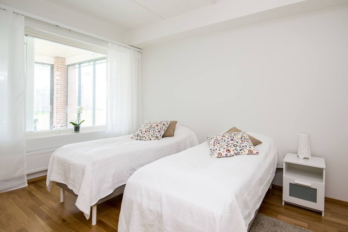 A bedroom with two single beds, a nightstand, a window with blinds, and a radiator.