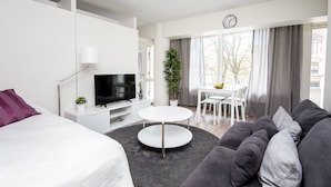 A modern hotel room with a bed, a TV, a dining area with a table and chairs, a sofa, and a large window with curtains.