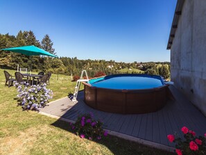 An outdoor swimming pool with a wooden deck, surrounded by a grassy area and a patio with outdoor furniture. - Beautiful holiday home near lake and forest (Savignac-Ledrier)