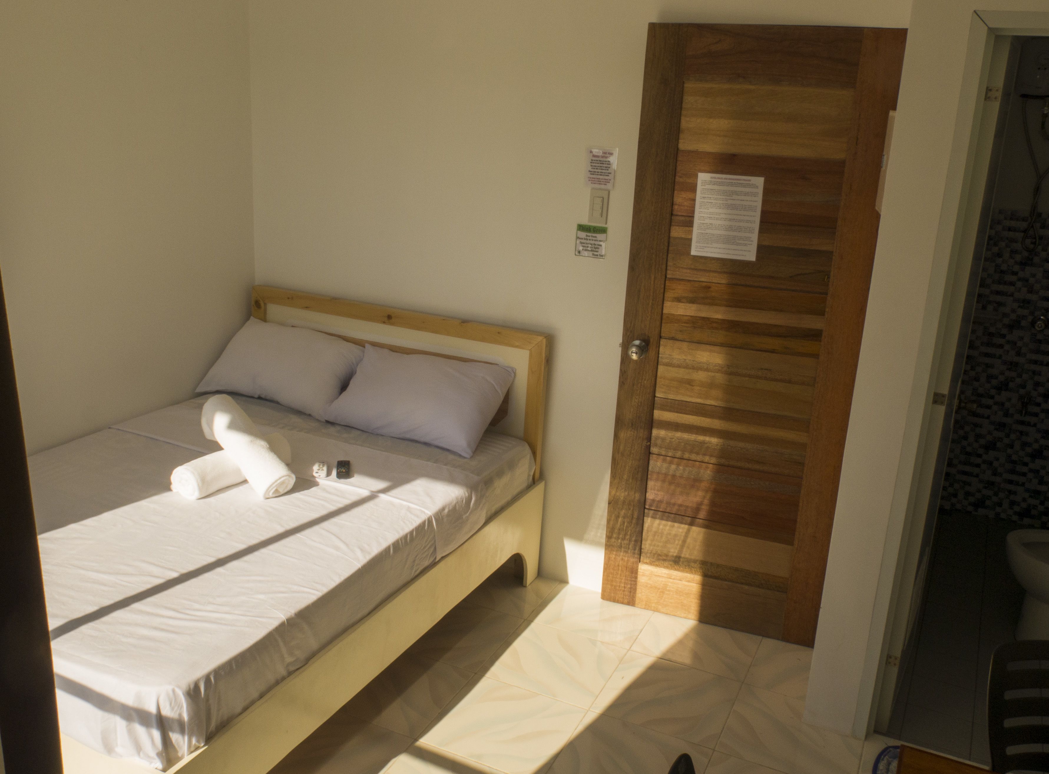Una cama individual con cabecera de madera, colchón blanco y dos almohadas. Hay una puerta de madera con un cartel y suelo de baldosas.
