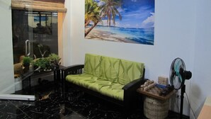 Une pièce avec un canapé vert, une porte en verre, un tableau représentant une plage et un ventilateur.