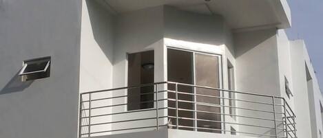 A corner of a building with a balcony, a window, and a small ventilation unit.
