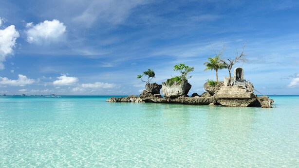 Une mer d’un bleu turquoise limpide, avec une île rocheuse parsemée de palmiers et sur laquelle se trouve une petite construction.