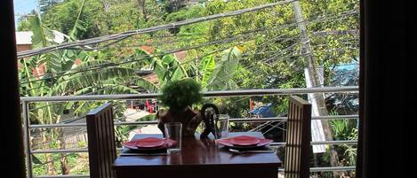 Une table à manger avec des chaises, dressée pour deux personnes, sur un balcon offrant une vue sur une colline et des lignes électriques.
